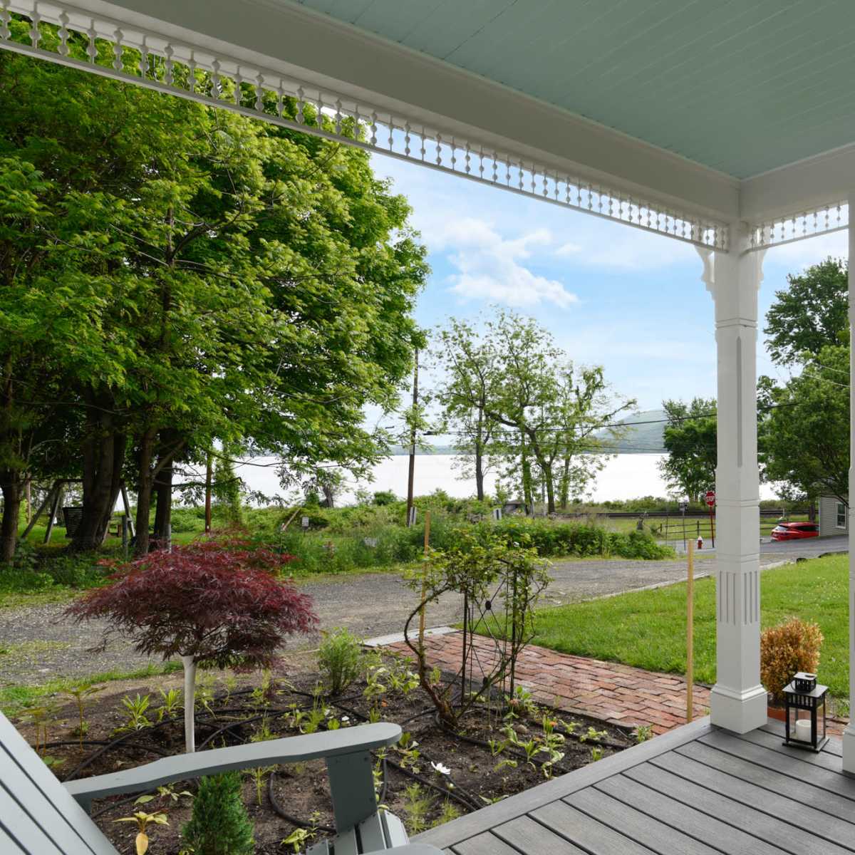 A covered porch offers a view of a green lawn, trees, and a distant lake under a blue sky.