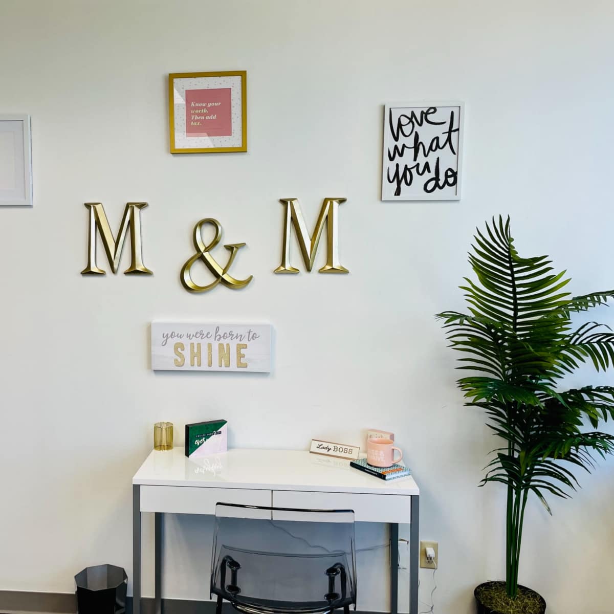 A minimalist workspace featuring a white desk, a clear chair, decorative wall art, and a small green plant in the corner.