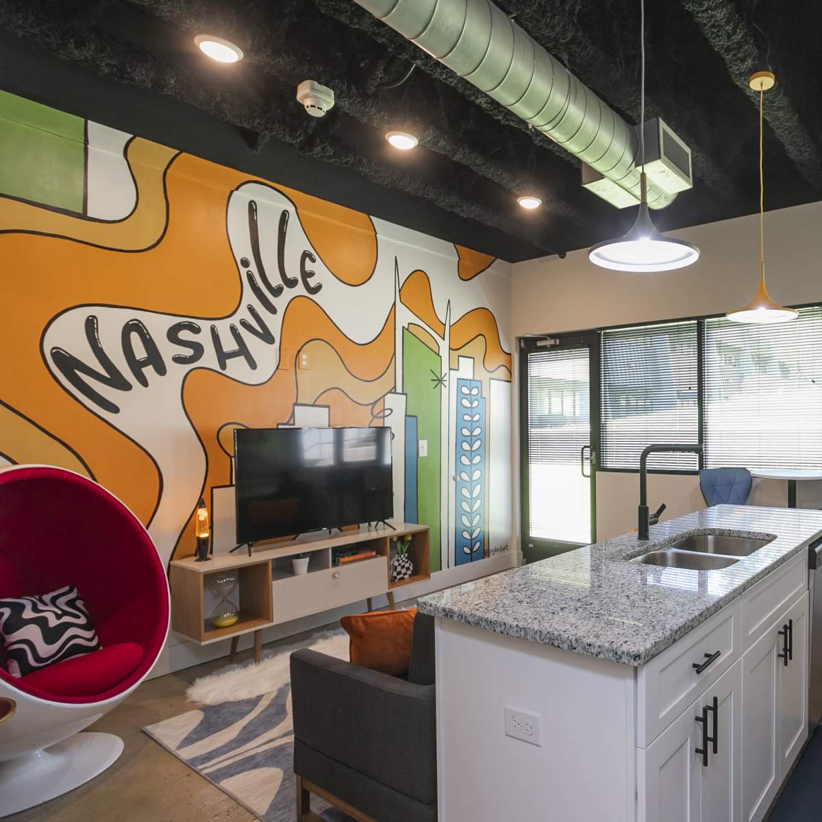 A modern kitchen and living area features a vibrant Nashville mural on the wall, with contemporary furnishings and large windows allowing natural light.