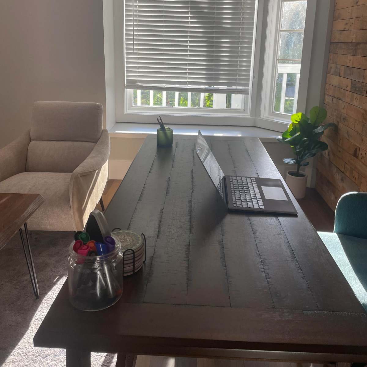 A modern office space features a wooden table with a laptop, a potted plant, and a chair, illuminated by natural light from a window.