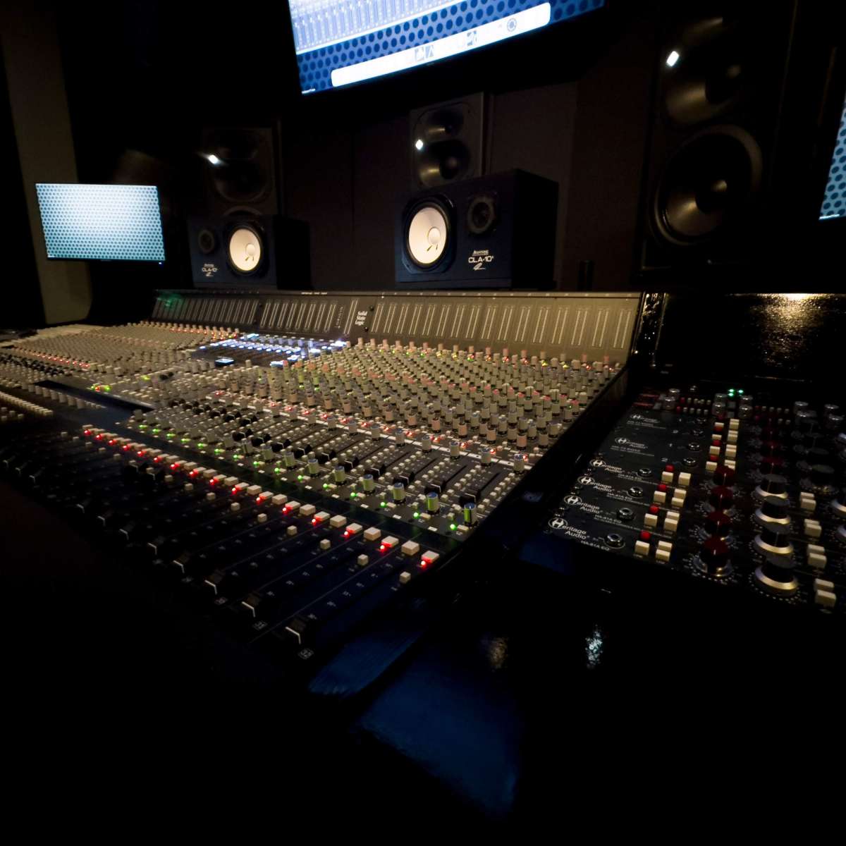 A music recording studio features a large mixing console surrounded by monitors and studio speakers.