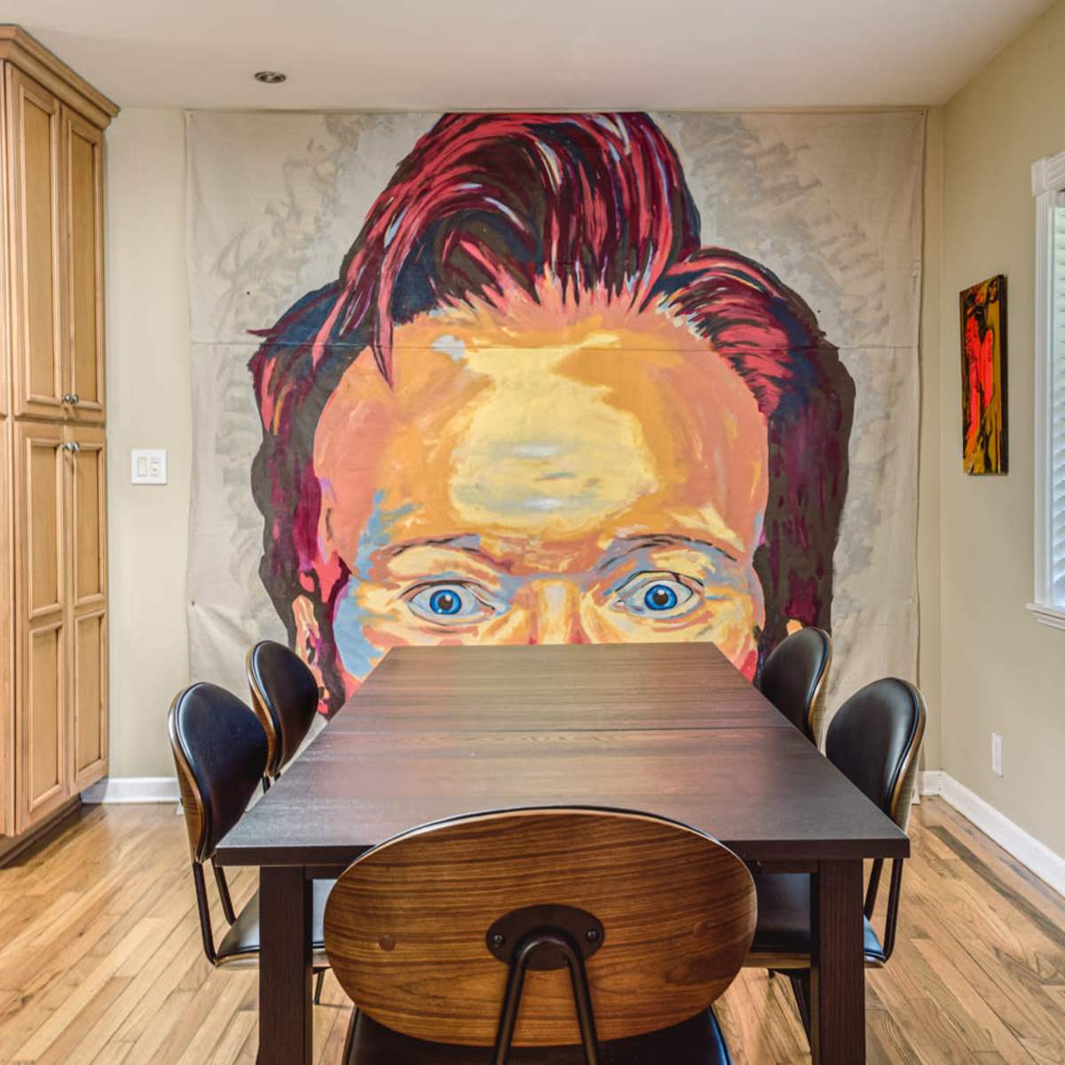 A large mural of a person's face dominates the wall above a modern dining table surrounded by black chairs in a well-lit kitchen.