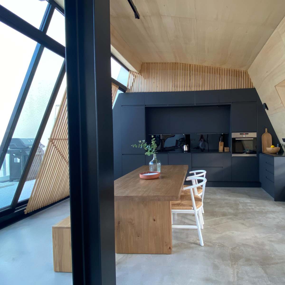 A modern kitchen with a wooden table and black cabinetry, featuring large windows and angled walls.