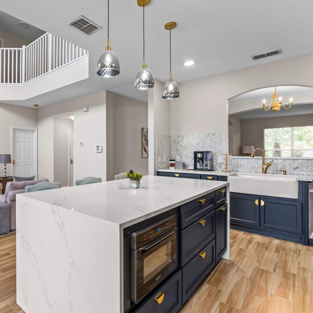 A modern kitchen with a large island, pendant lighting, and a living area in the background, showcasing a light color scheme and comfortable seating.