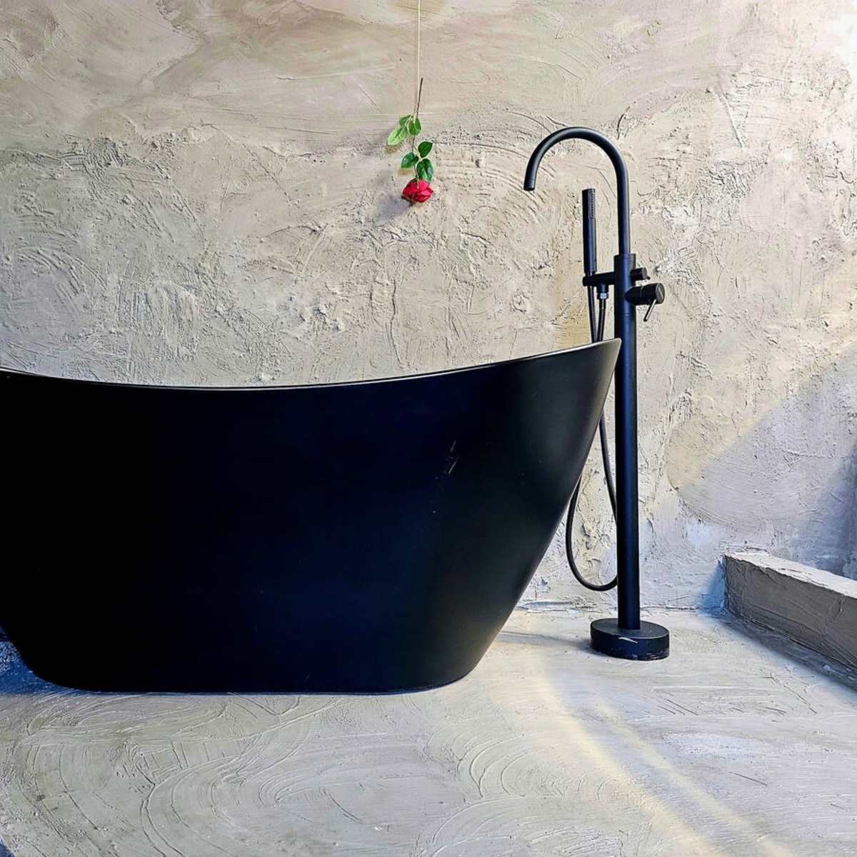 A modern black bathtub on a textured concrete surface, accompanied by a floor-mounted faucet and greenery in the background.