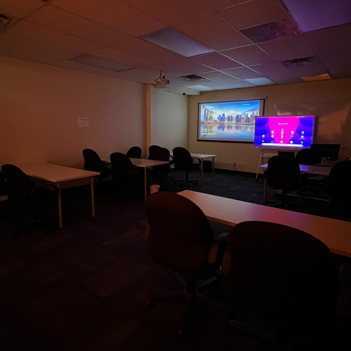 A dimly lit classroom features several empty tables and chairs, with a projector screen and a television displaying a digital interface on one wall.