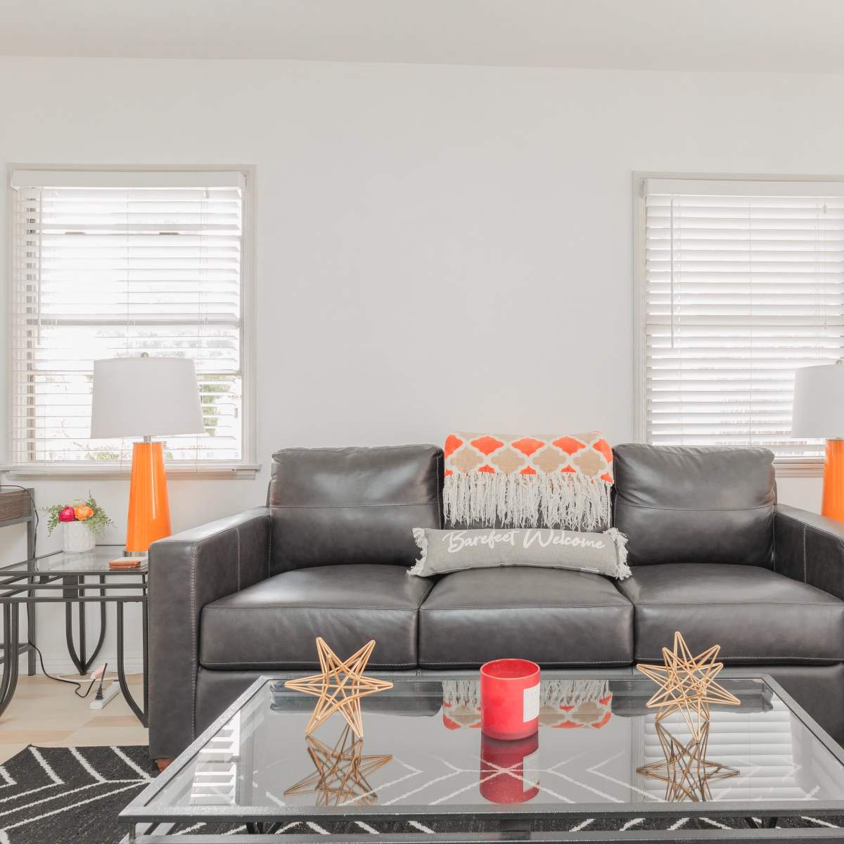 A modern living room features a black leather sofa with a decorative throw, two orange lamp tables, and a coffee table adorned with star-shaped decor.