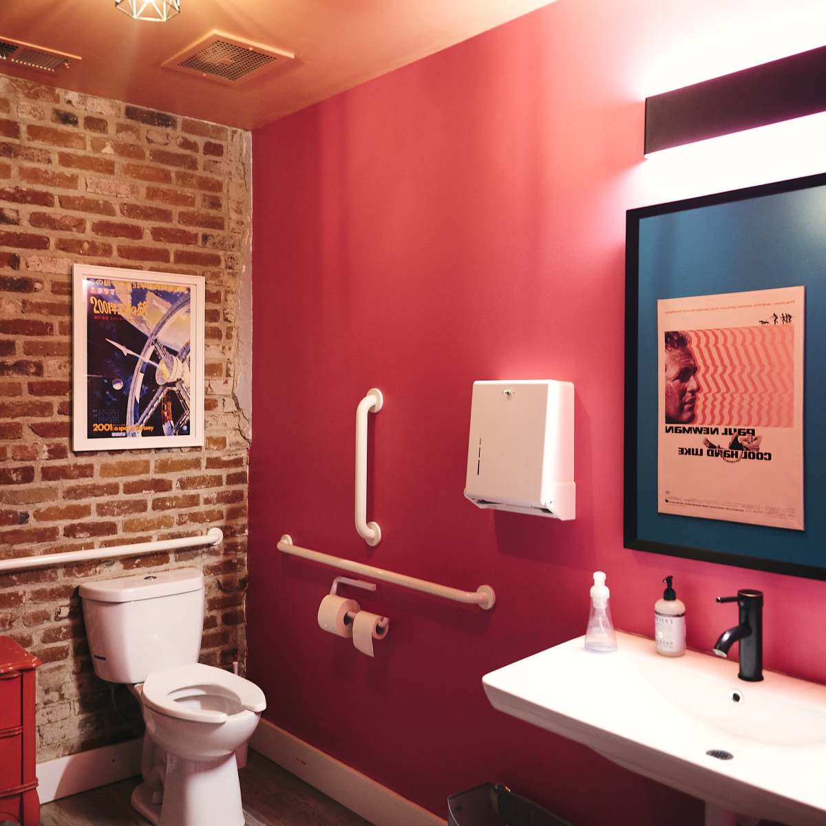 A modern bathroom featuring a toilet, a wall-mounted sink, a colorful poster, and a brick wall with exposed sections.