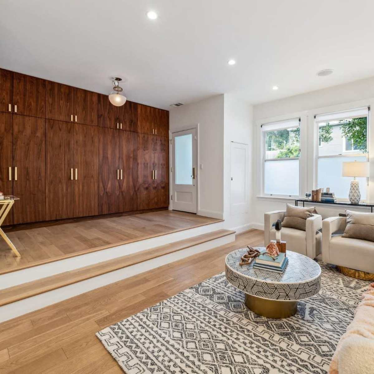 A modern living room featuring a neutral color palette, a raised wooden area with built-in cabinetry, and large windows allowing natural light.