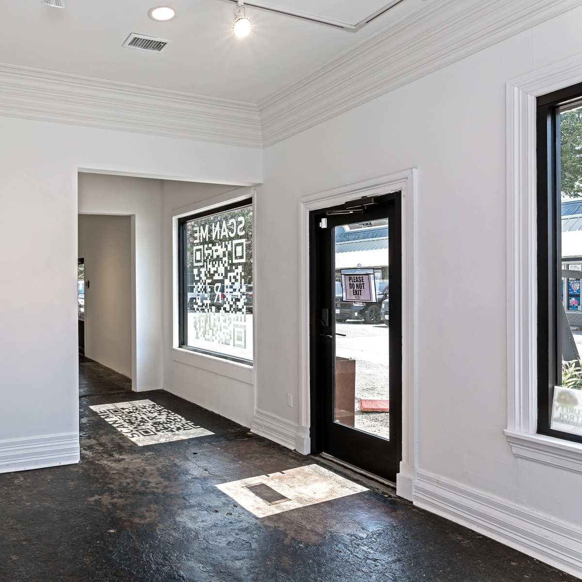 A well-lit, empty interior space with white walls, dark flooring, and a large window facing the street.