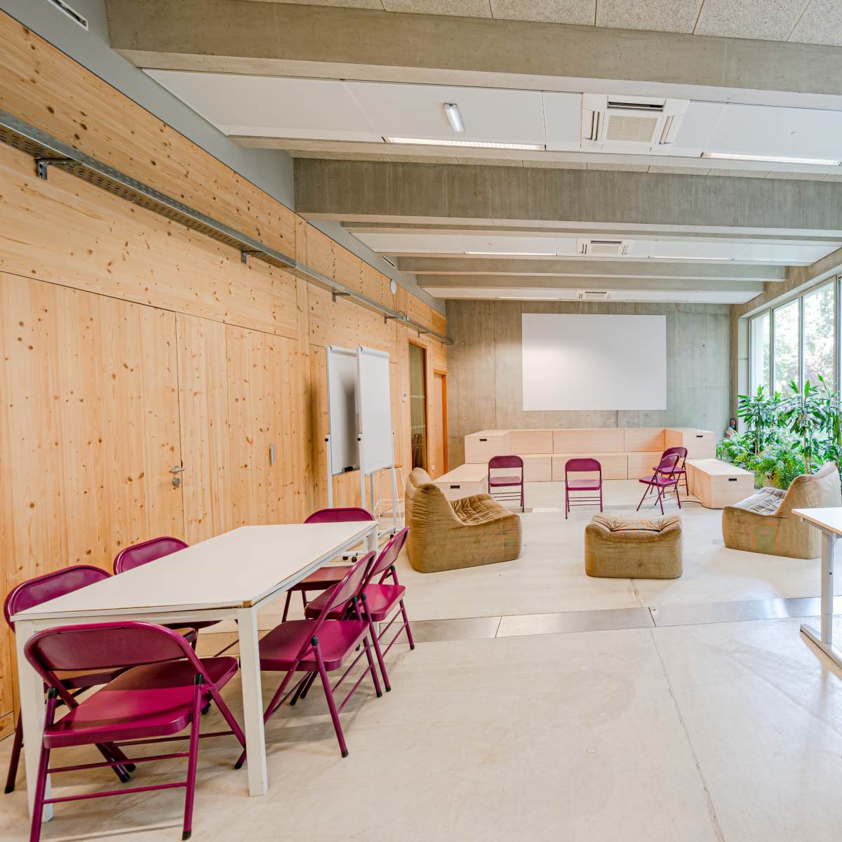 A modern classroom with several tables and chairs arranged around a central seating area, surrounded by large windows and greenery outside.