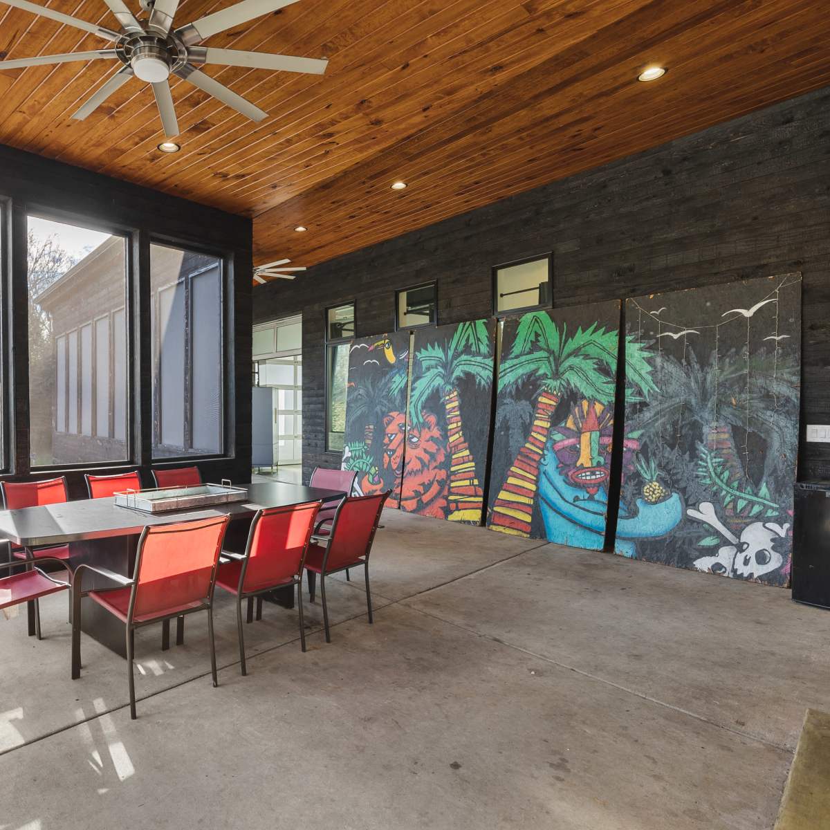 A spacious indoor dining area with a large table and red chairs, featuring large windows and a colorful mural on one wall.