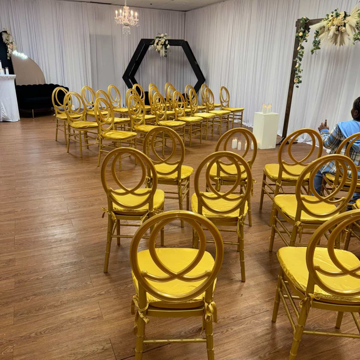 A room set up for an event, featuring rows of yellow chairs arranged in front of a black hexagonal arch and decorated with floral elements.