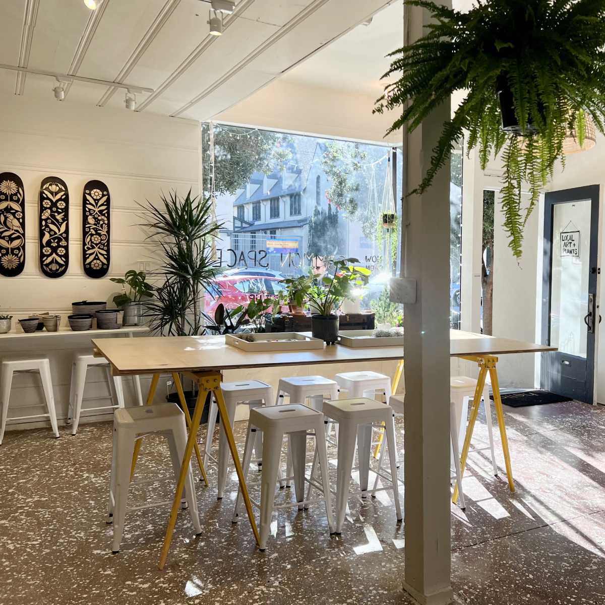 A bright interior of a contemporary space with a long wooden table surrounded by white stools, plants, and large windows allowing natural light.
