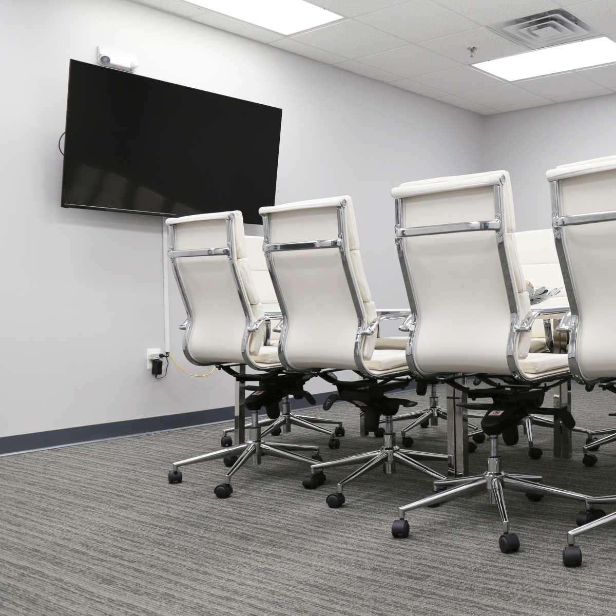 A conference room with a long table and several white office chairs arranged around it, and a wall-mounted TV at one end.