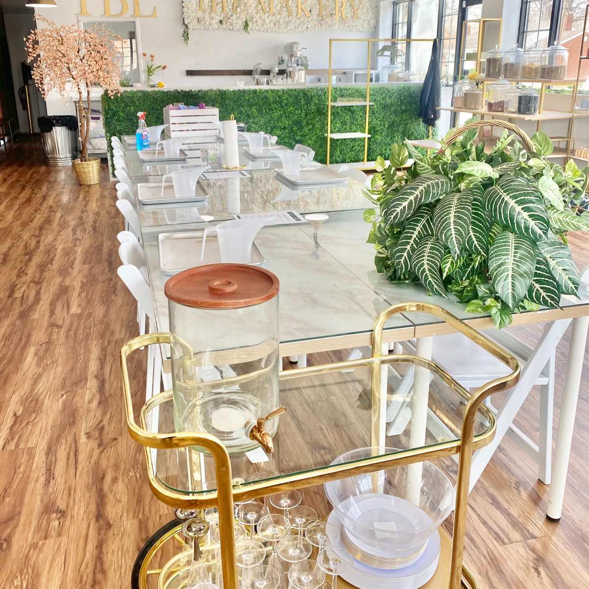 A bright, modern bakery interior with a long table, white chairs, and decorative plants, featuring a cart with glassware and a water dispenser.