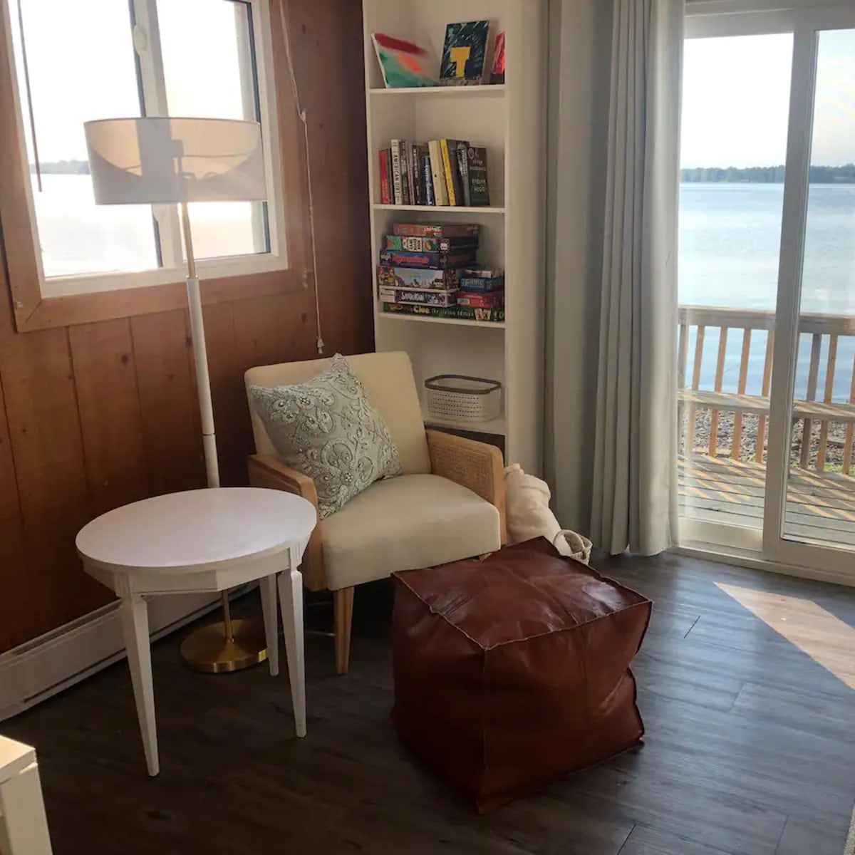 A cozy corner of a room features a light-colored armchair, a round white table, a brown pouf, and a large window overlooking a calm lake.