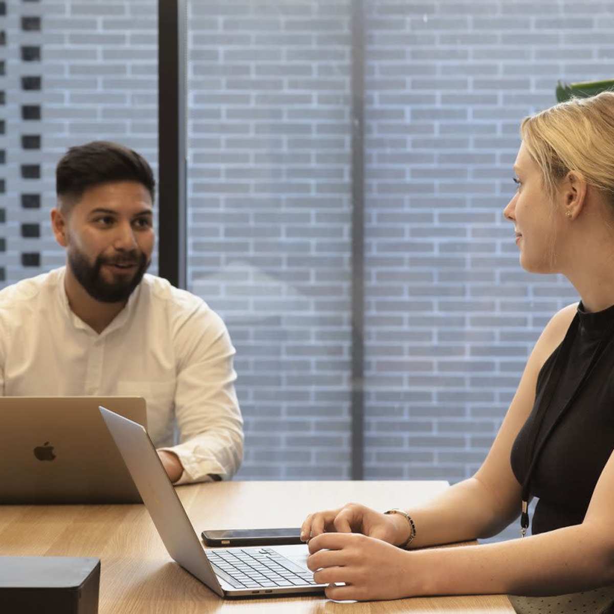 Two professionals are sitting at a wooden table in a meeting room, one using a laptop while the other has a laptop open in front of them, with a large window and a plant visible in the background.