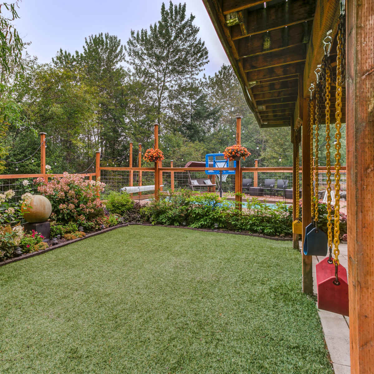 A landscaped backyard with synthetic grass, colorful flower beds, a wooden deck, and a swing set.