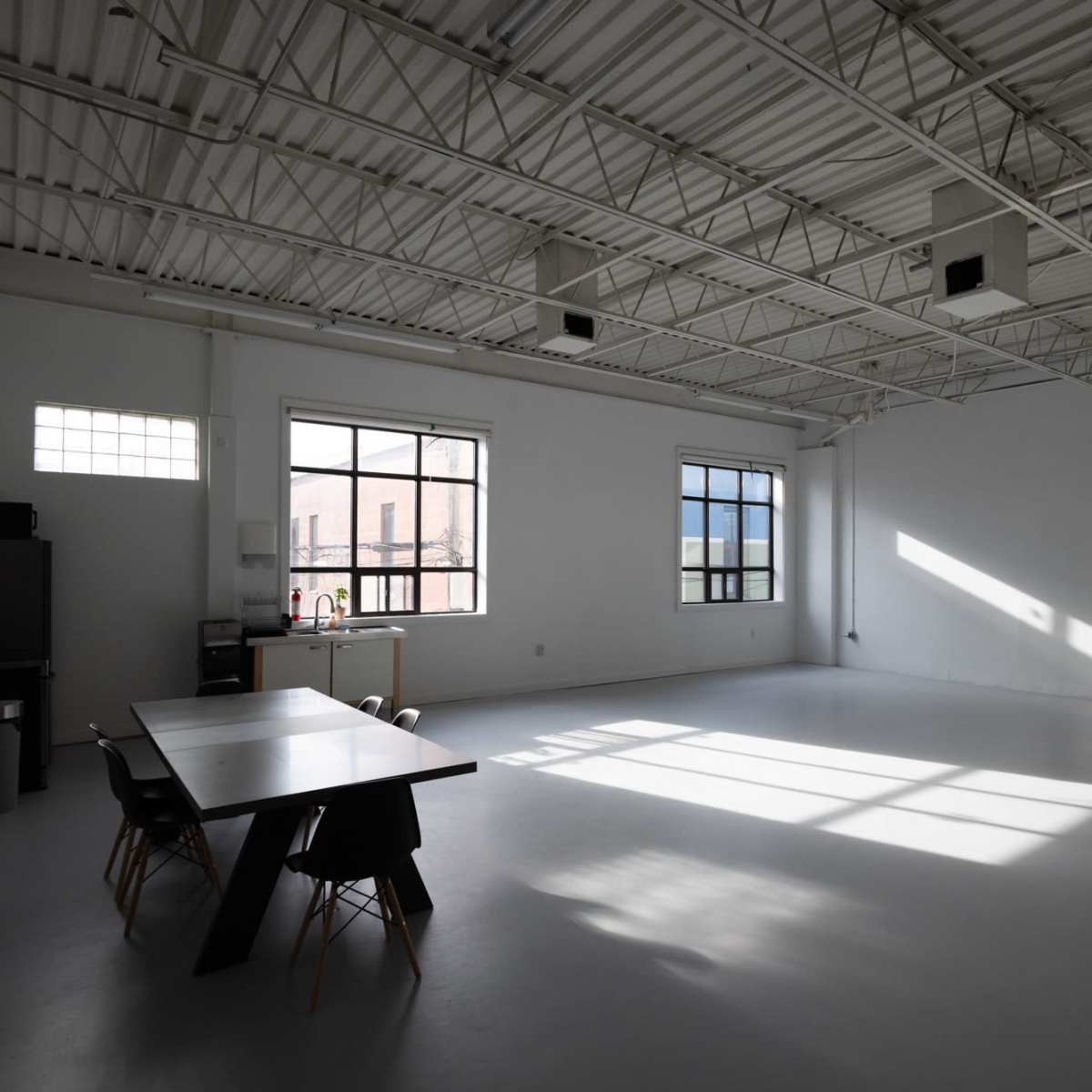 A large, empty room with white walls, a long table, and several chairs, illuminated by sunlight streaming through the windows.