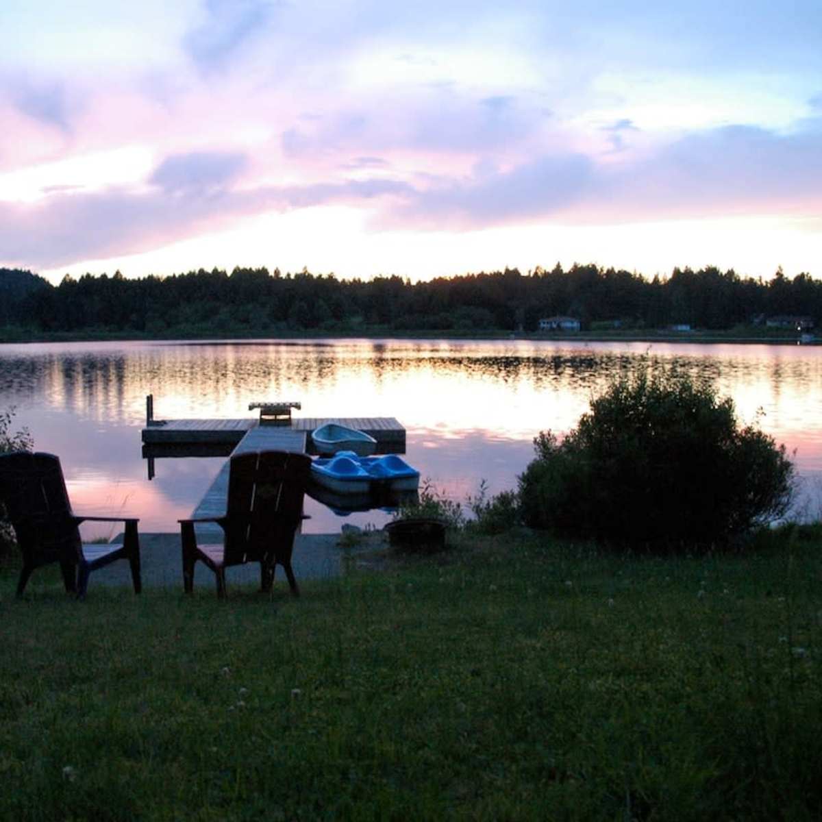 Two chairs sit on the grass by a calm lake, with a dock and a blue boat visible in the water beneath a colorful sky at sunset.