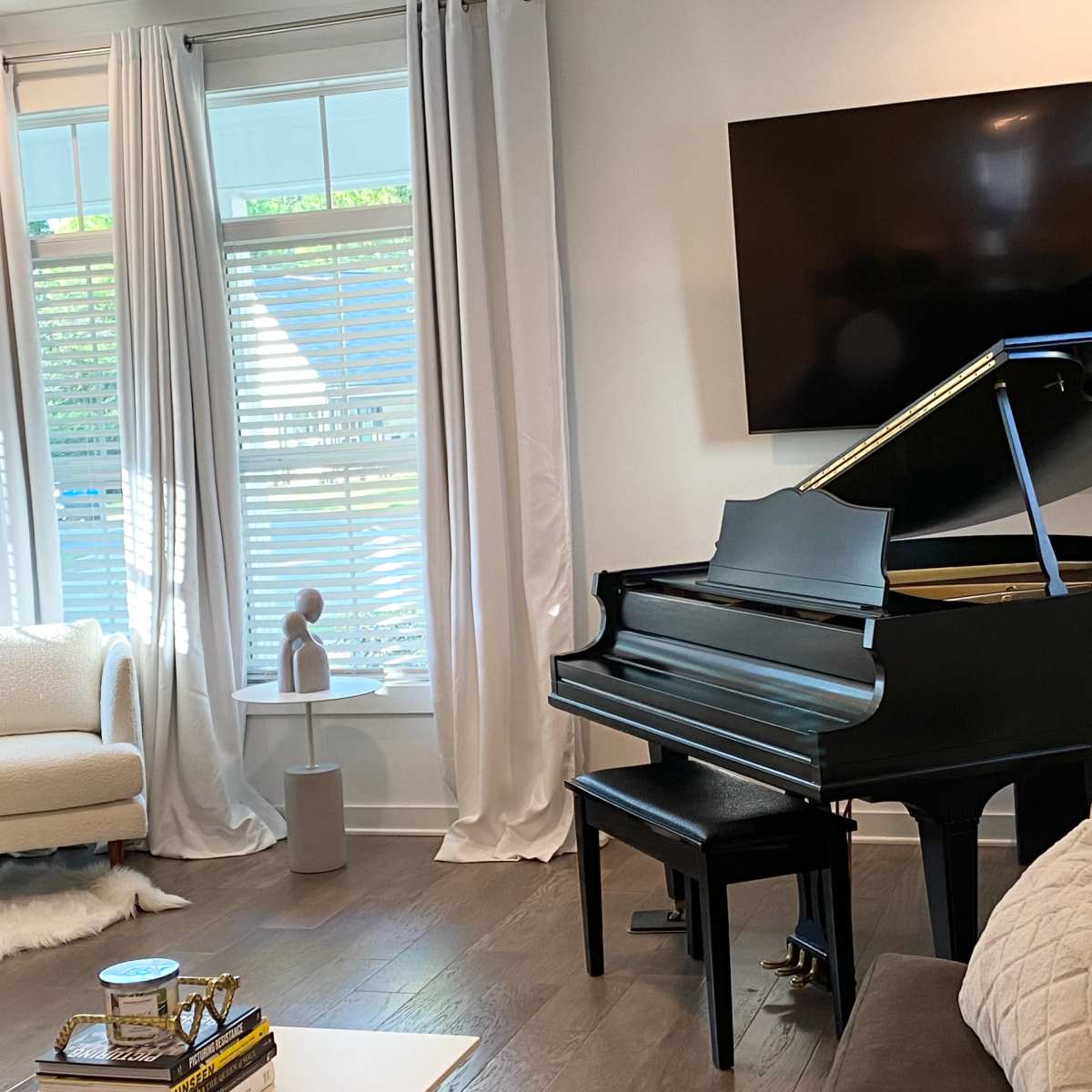 A dark grand piano is positioned beside a modern chair in a bright living room with large windows and a mounted television.