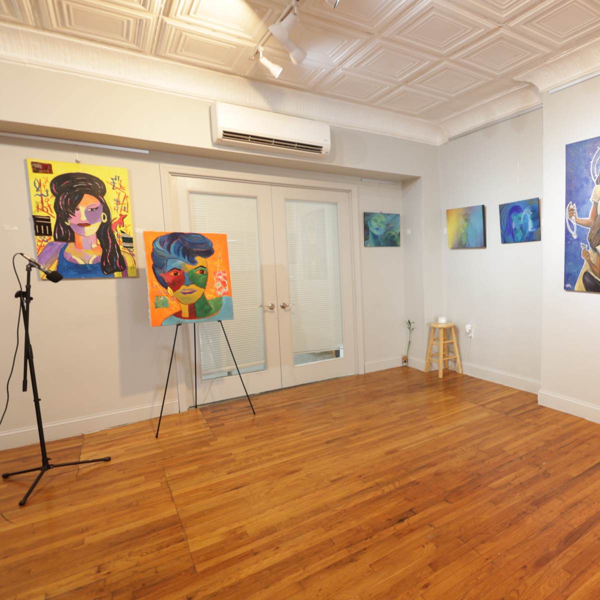 An art gallery with colorful paintings displayed on the walls, a microphone stand positioned in the corner, and wooden flooring.