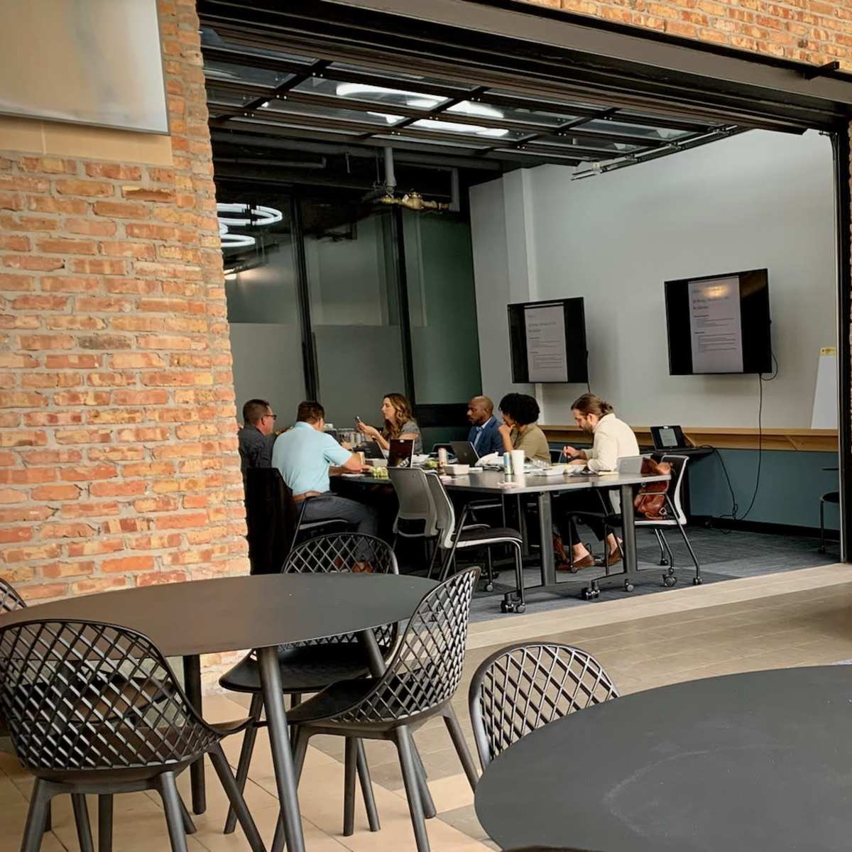 A group of people is seated around a table in a meeting room, while others relax on sofas in an open office space with brick walls.