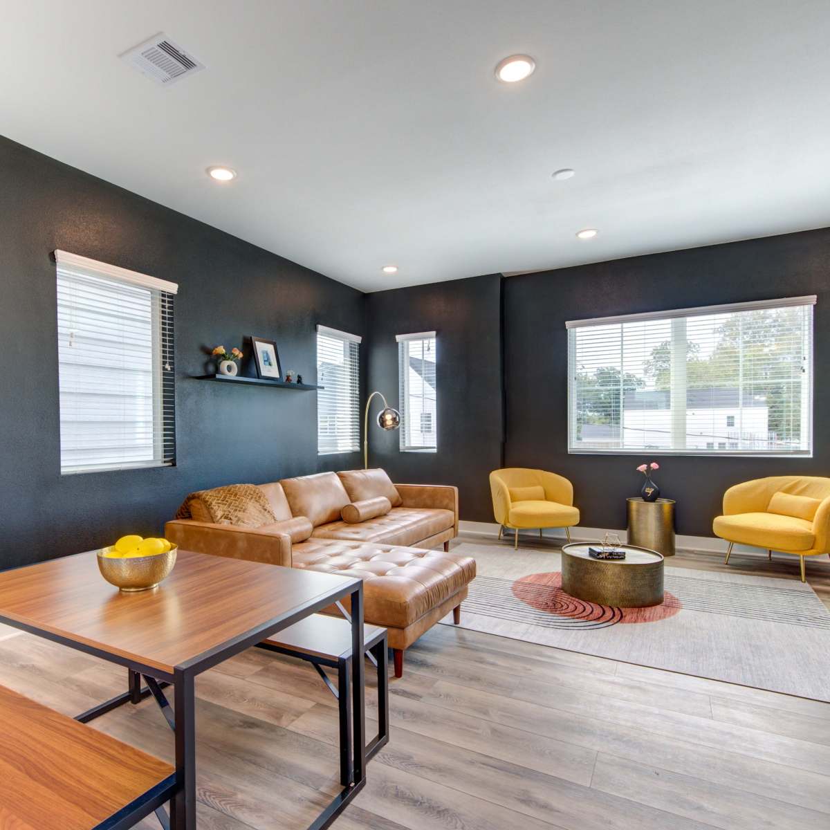 A modern living space featuring a brown leather sofa, two yellow armchairs, a wooden table, and a round coffee table on a light rug against dark walls.