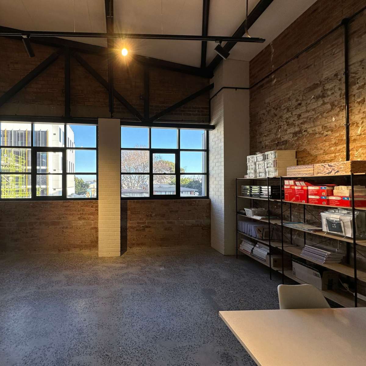 A spacious room with exposed brick walls, large windows, and a shelf filled with boxes and files.