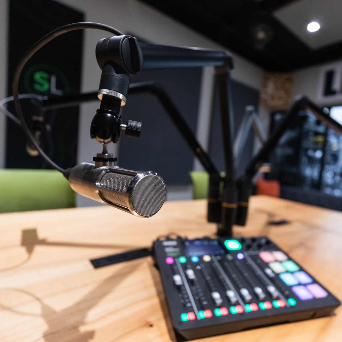 A microphone is positioned on an adjustable arm above a mixing console with colorful buttons and sliders on a wooden table in a recording studio.