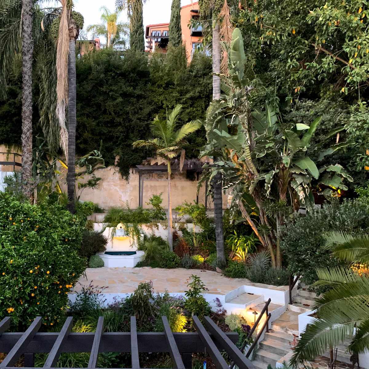 A landscaped garden with various plants and palm trees, featuring a small fountain at the center surrounded by stone pathways.