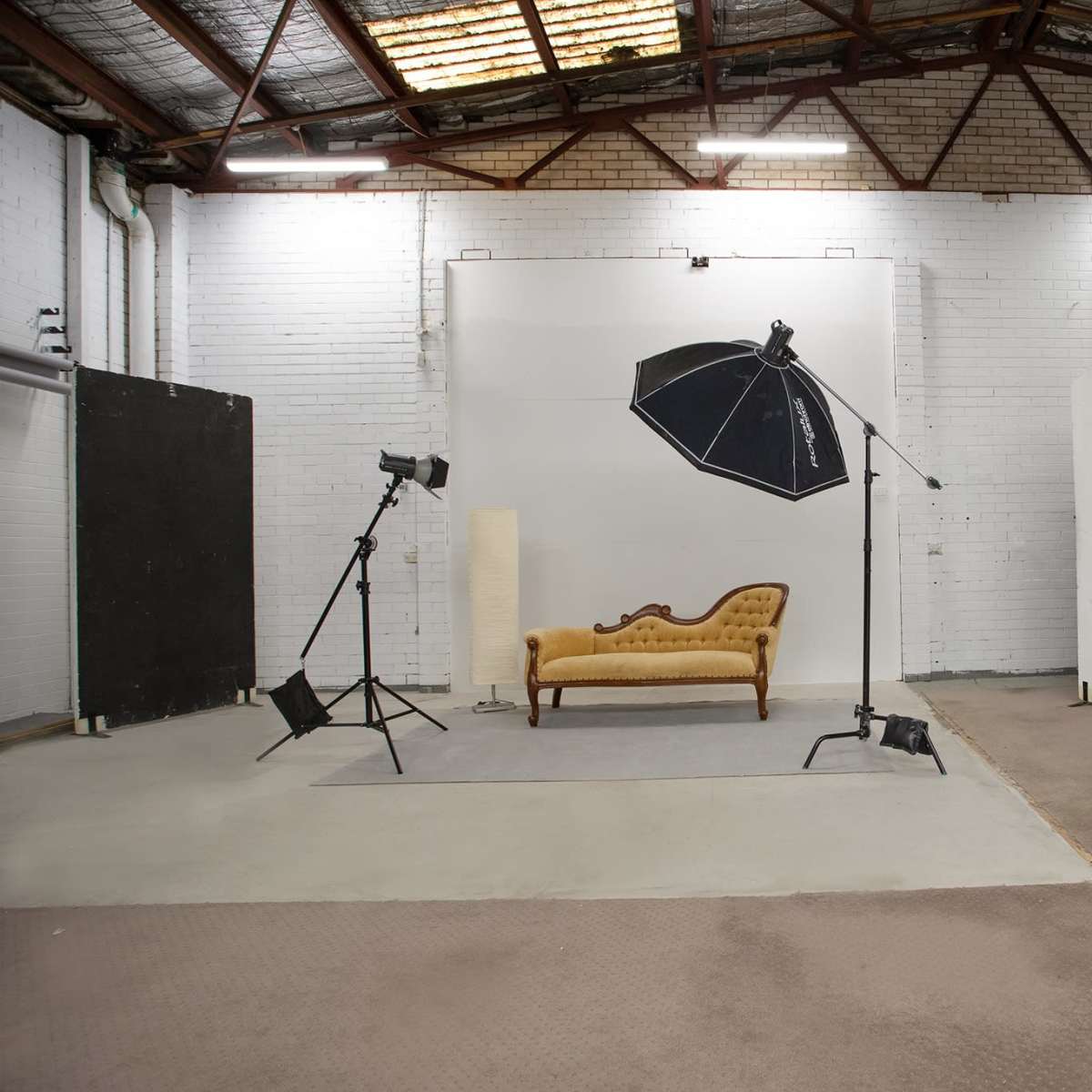 A photo studio featuring a vintage-style couch, a large umbrella light, and various backdrops in a spacious, industrial setting.