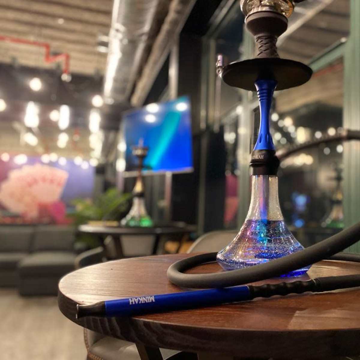 A hookah with a blue base and black hose is placed on a wooden table in a lounge setting with soft seating and a television in the background.