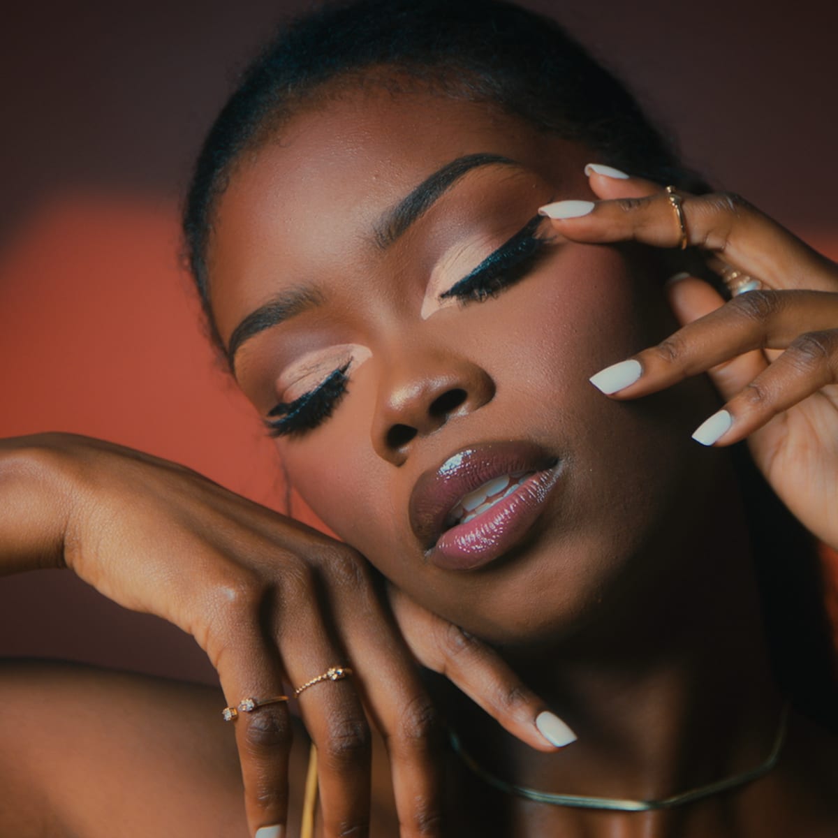 A woman poses gracefully with her hands framing her face against a softly lit background.