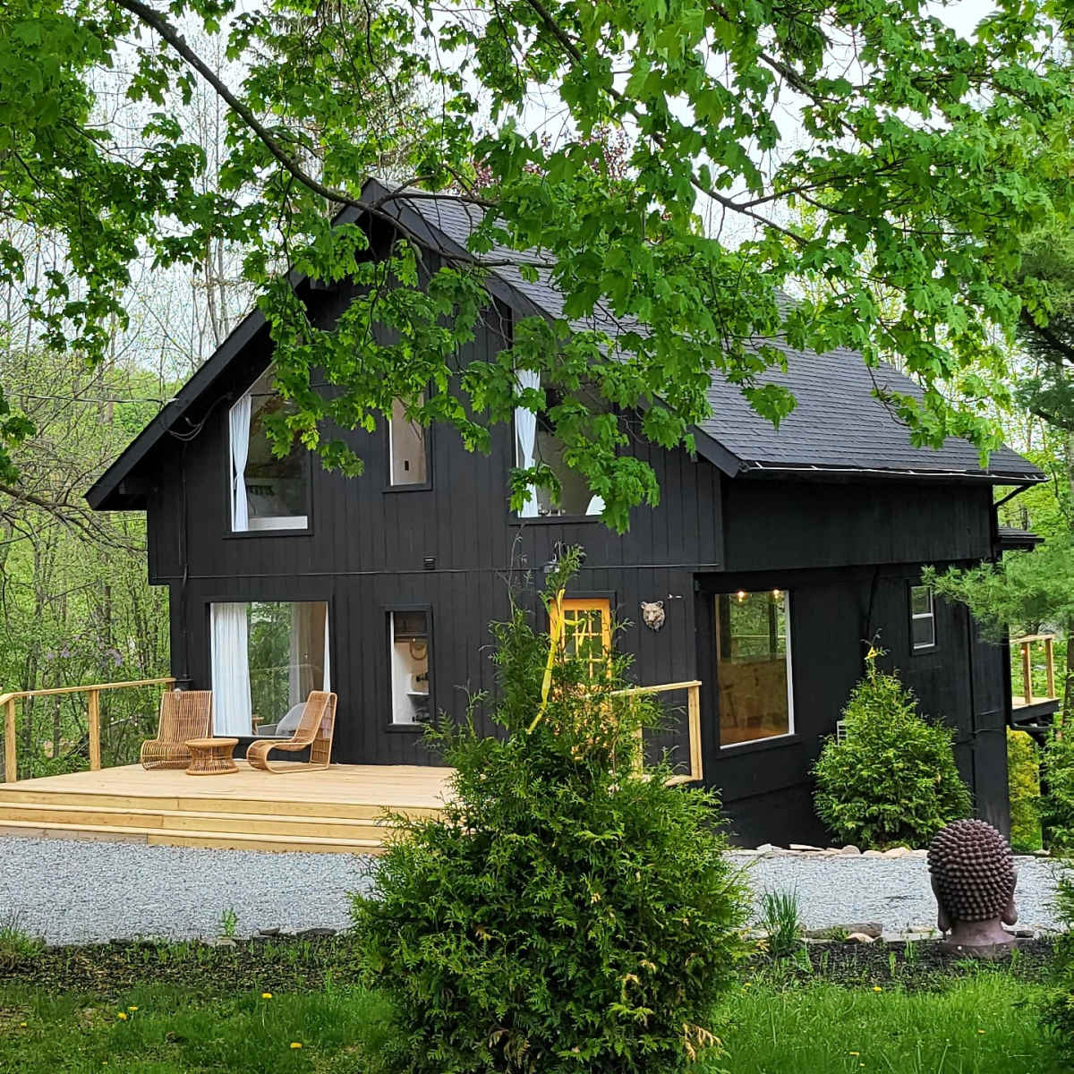 A dark-colored house surrounded by greenery, set back from the road with a wooden deck and large windows.