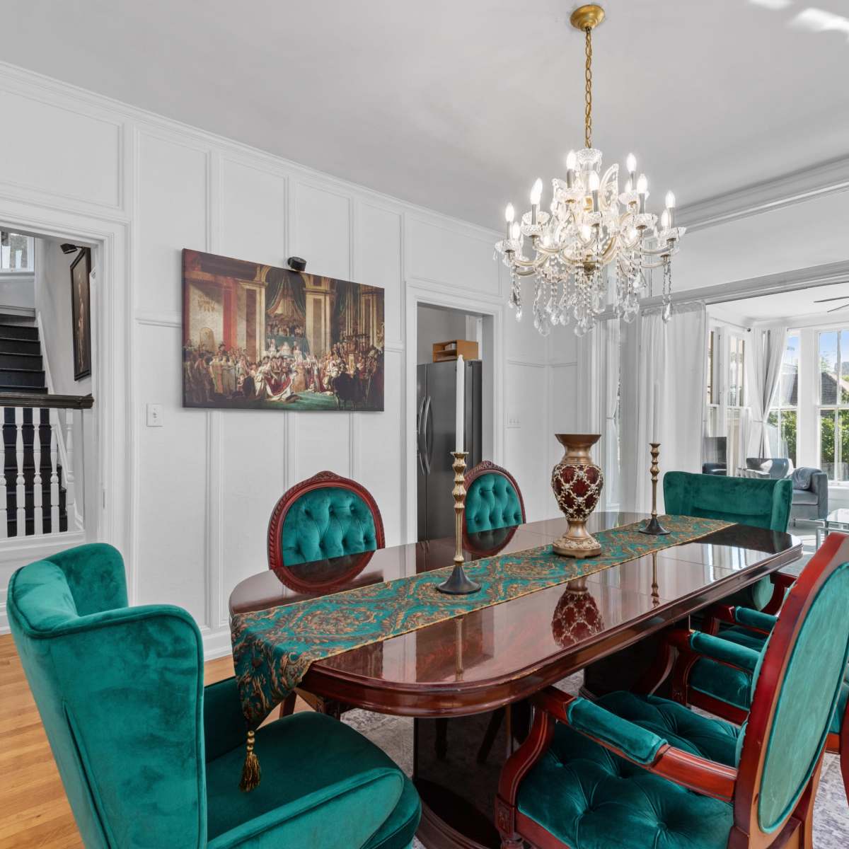 A dining room features a polished wooden table surrounded by green velvet chairs and a chandelier overhead, with large windows allowing natural light to enter.