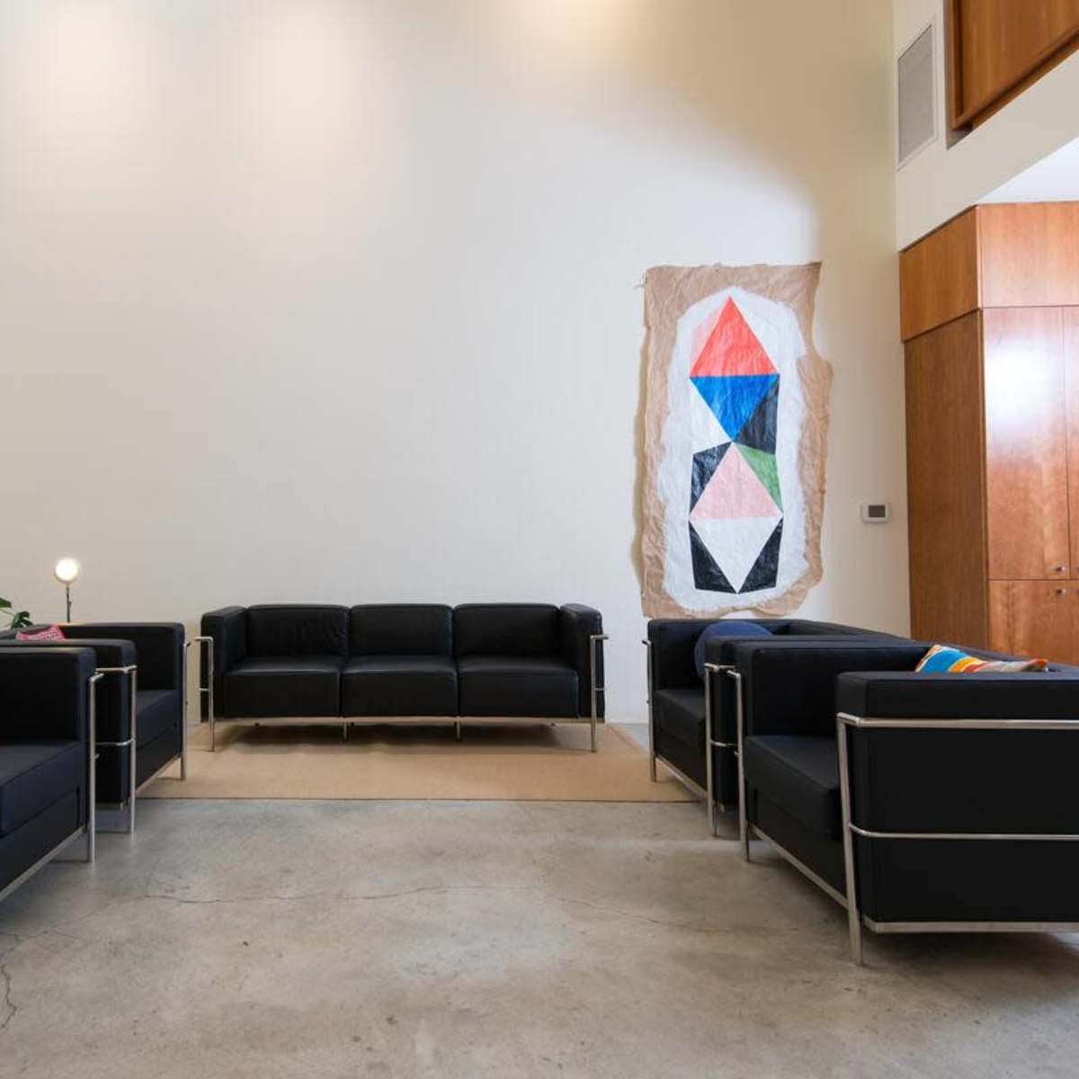 A modern living area with four black sofas arranged around a central space, a geometric wall hanging, and wood cabinetry in the background.