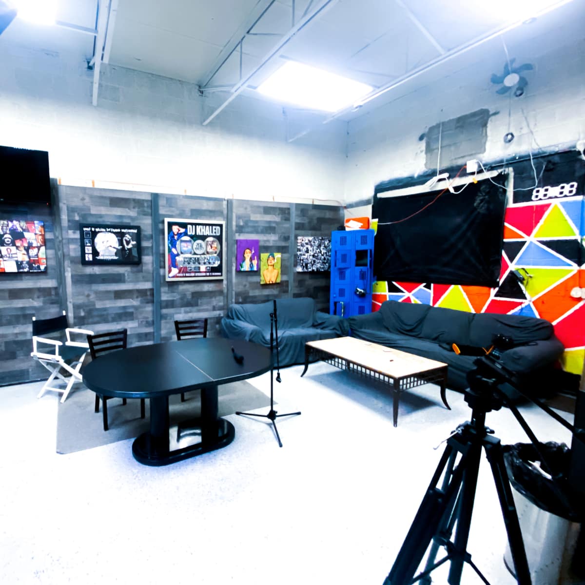 A studio space with a black table surrounded by chairs, colorful wall decorations, and camera equipment set up for recording.