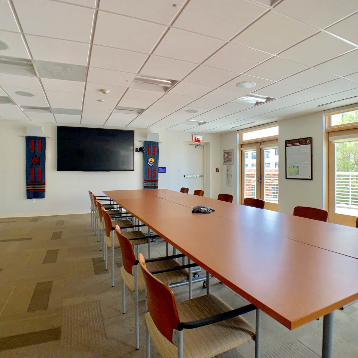 A spacious conference room features a long wooden table, chairs, a large wall-mounted screen, and windows that open to an outdoor view.