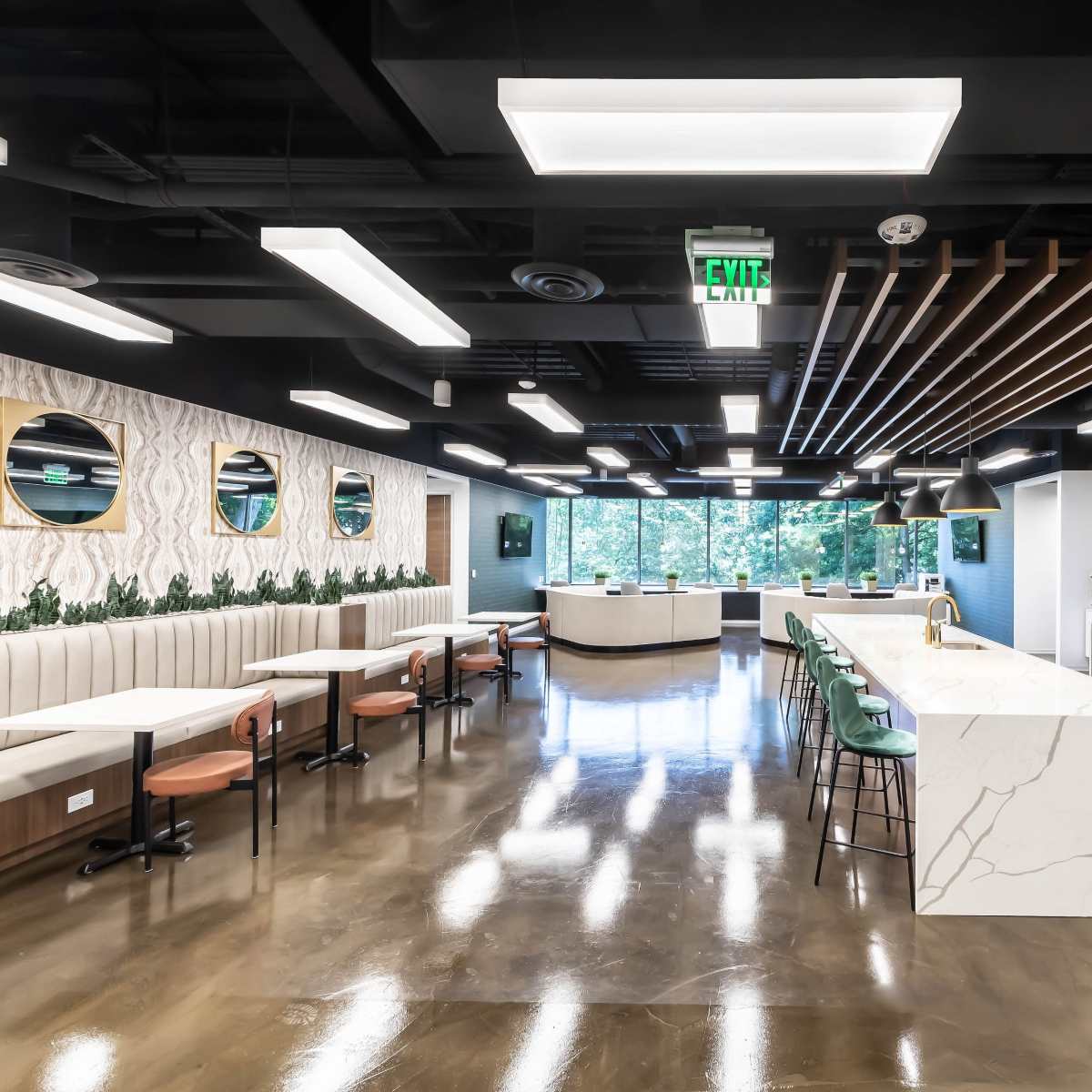A modern office kitchen area featuring a long marble countertop, seating along the walls, and large windows that provide ample natural light.