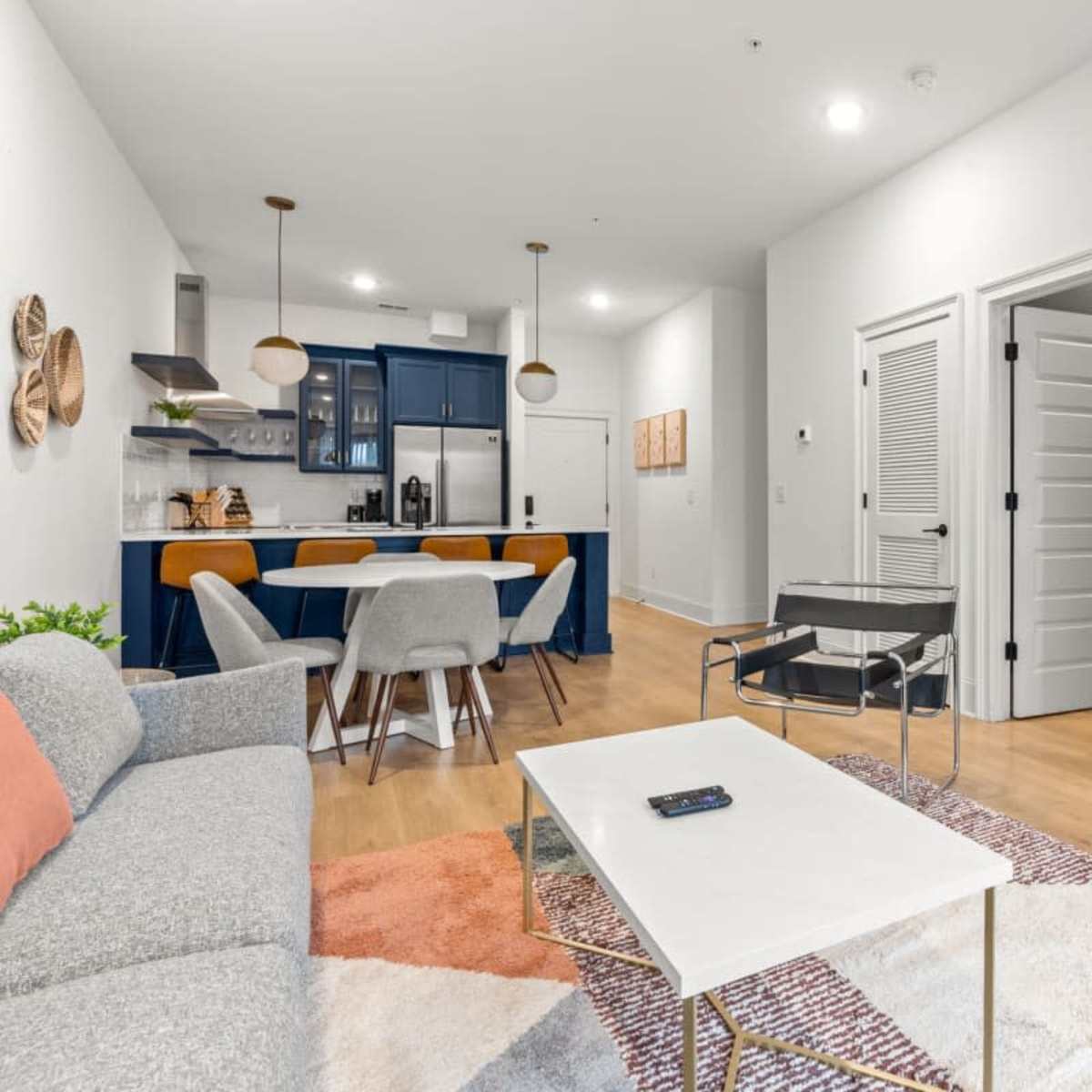 A modern living room with a gray sofa, a coffee table, and a TV mounted on the wall, leading into an adjacent kitchen area with blue cabinets.
