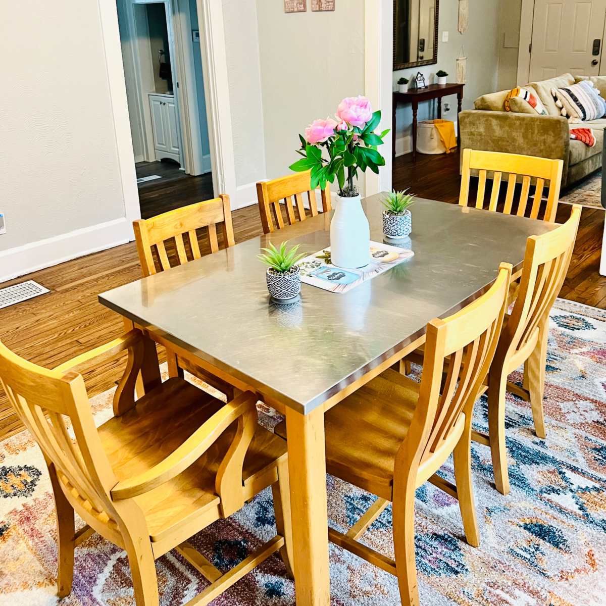 A modern dining room features a metal table surrounded by wooden chairs, with a vase of flowers and small plants on the table, and a cozy living area visible in the background.