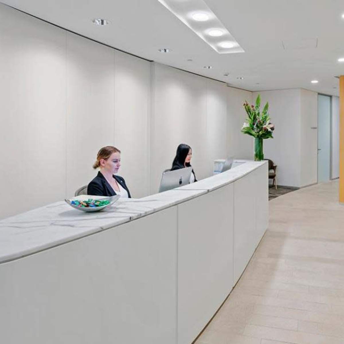 A modern reception area with a curved white desk, two seated women working, and minimalist furniture in a bright, open space.