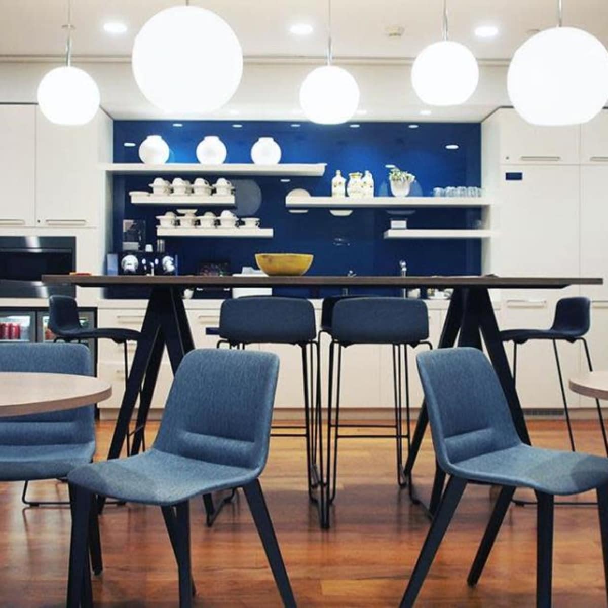 A modern kitchenette area features a long table with stools, surrounded by circular pendant lights and shelves displaying dishware.