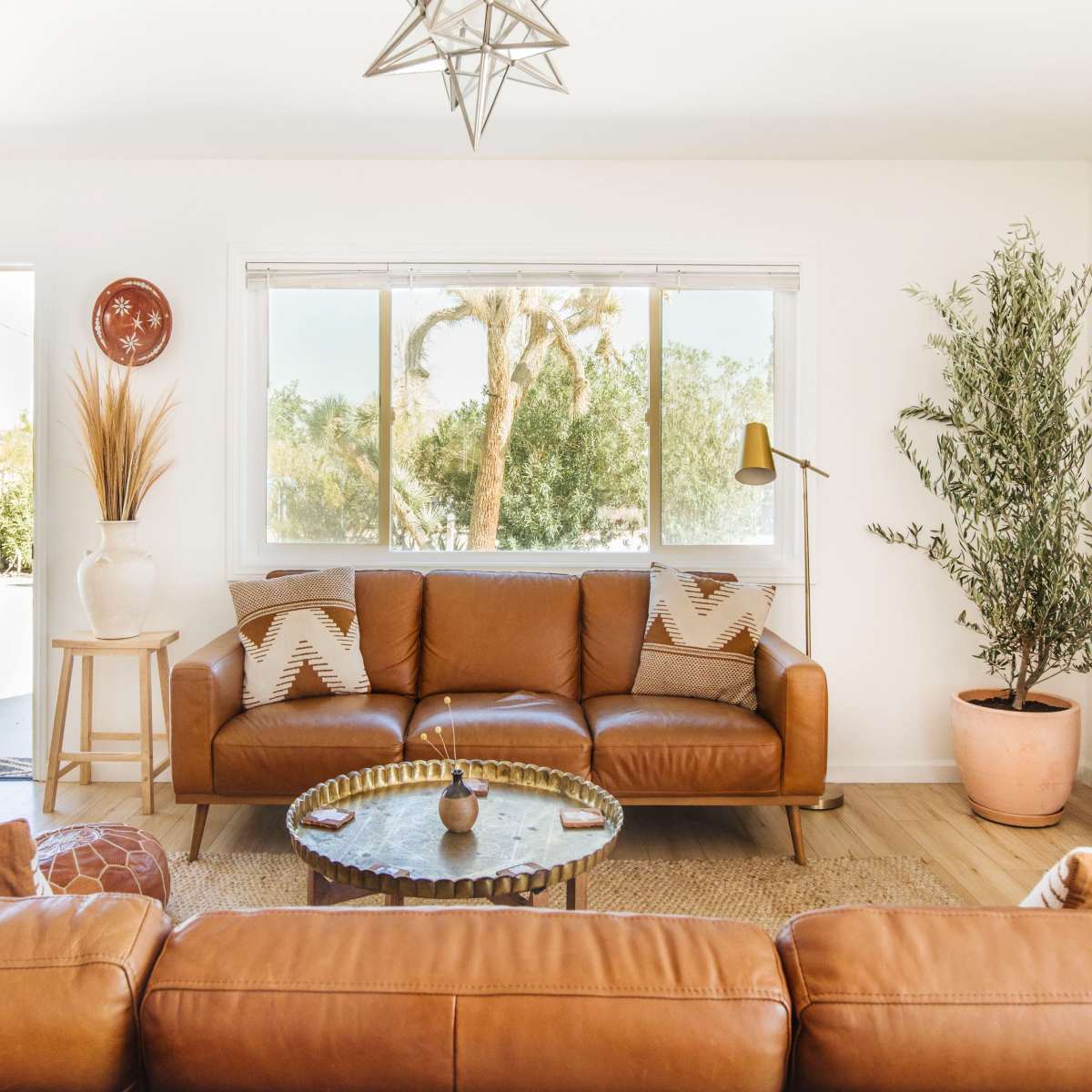A modern living room features a brown leather sofa, a round coffee table, and decorative plants, with a door leading to an outdoor space.