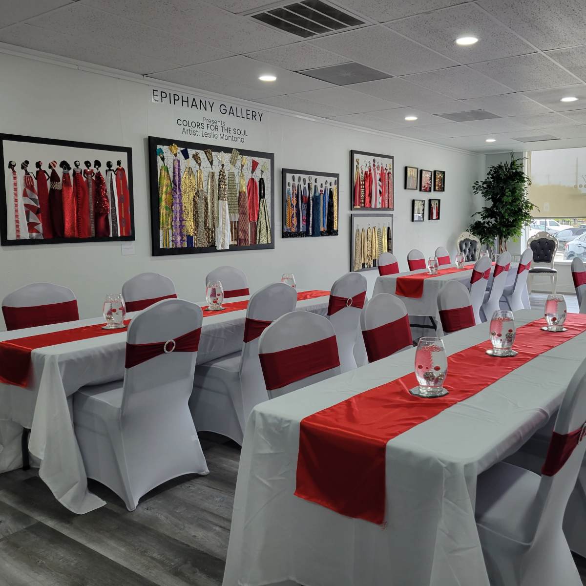 A well-decorated event space with tables covered in white cloths and red runners, surrounded by framed artwork on the walls.