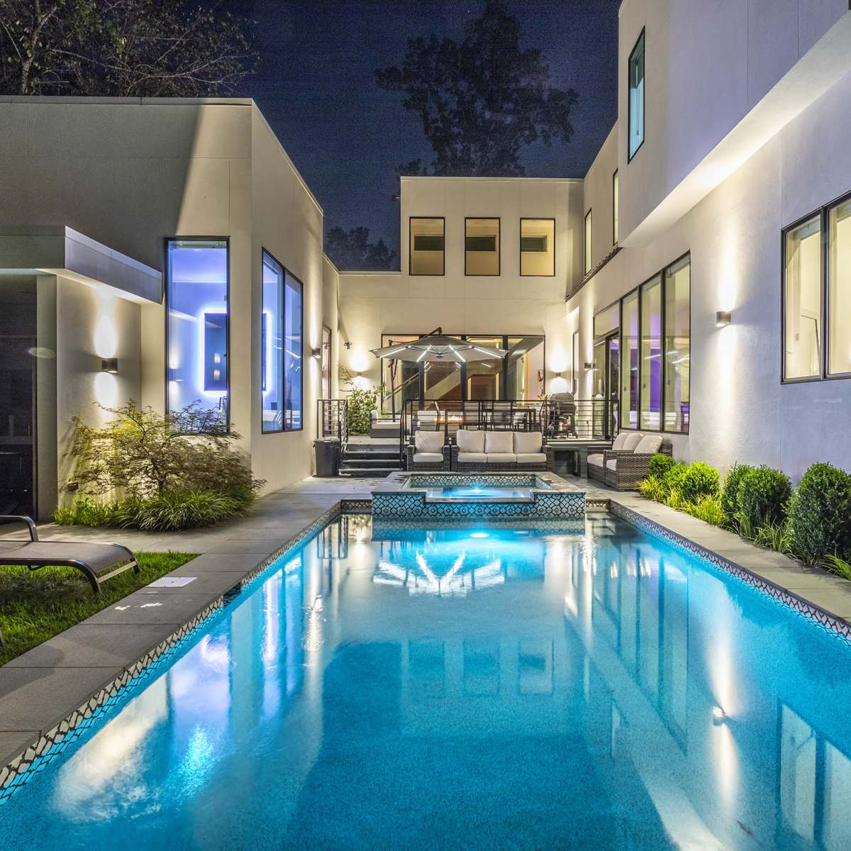 A modern home with large windows surrounds a illuminated swimming pool in a landscaped courtyard at night.