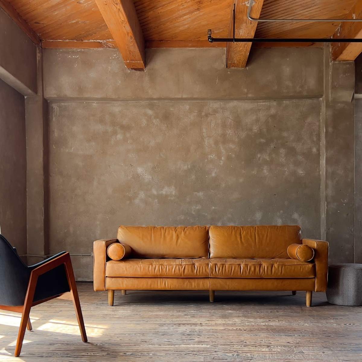 The room features a leather sofa, a modern chair, and large windows allowing natural light to illuminate the wooden floor and textured walls.