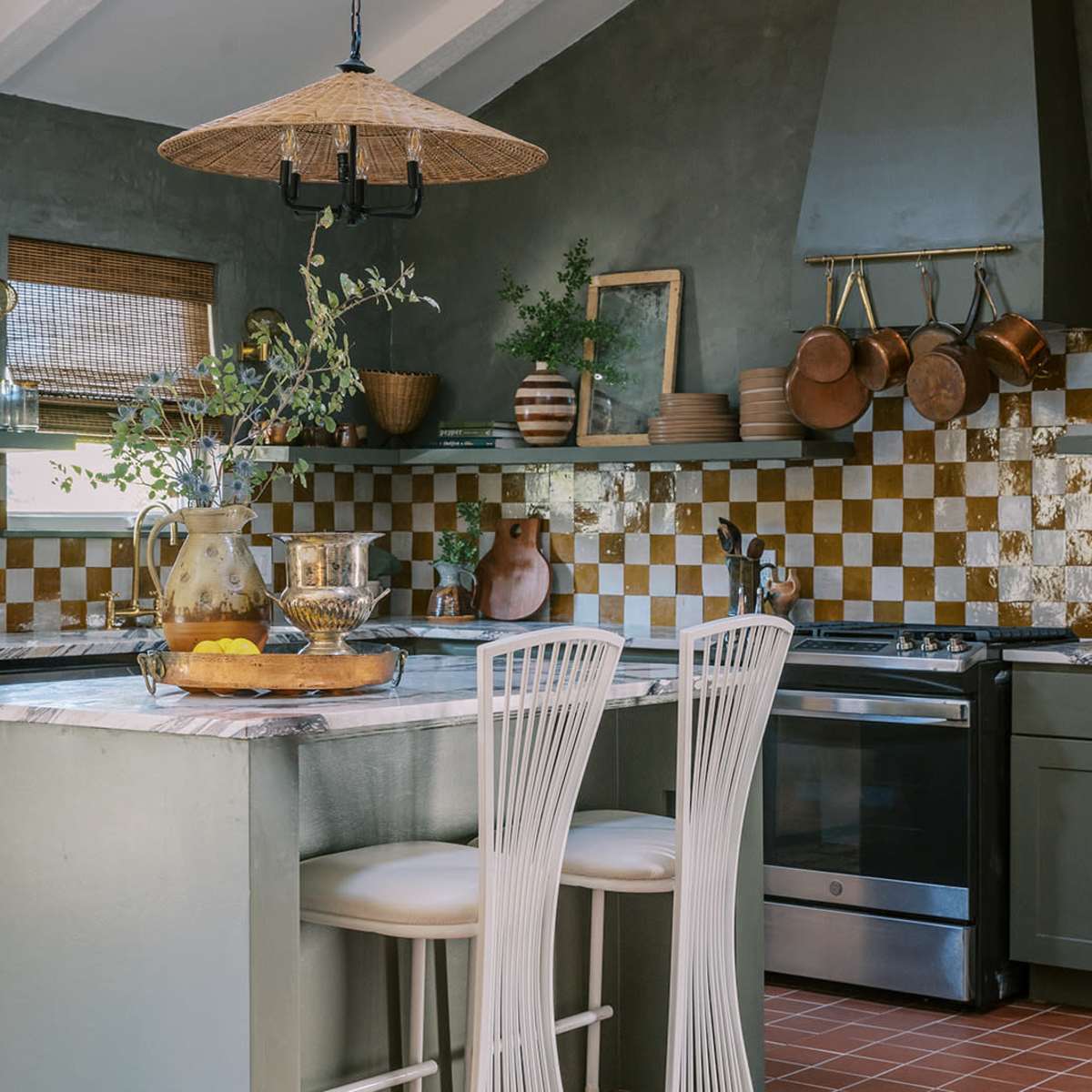 A kitchen with green cabinets, a marble countertop island, and a patterned backsplash in gold and white tiles, featuring hanging pots and decorative items.